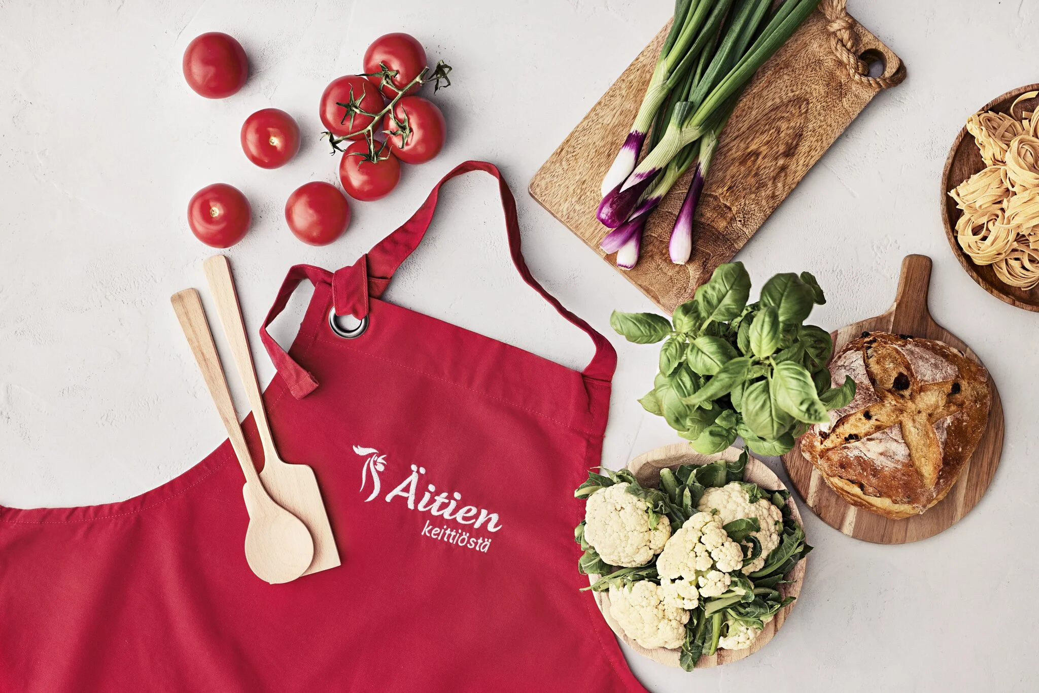 Kuvassa on essu, puukauha, tomaatteja, kukkakaalia, basilikaa, kevätsipulia, leipä ja pastaa.