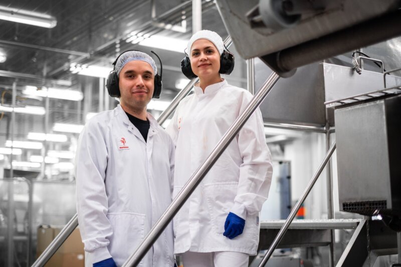 Kuvassa on kaksi henkilöä, jotka ovat suojavarusteisiin pukeutuneina tehtaalla.