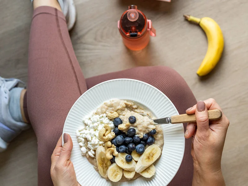 Kuvassa on henkilö urheiluvarustuksessa istumassa lattialla ja syömässä puuroa.