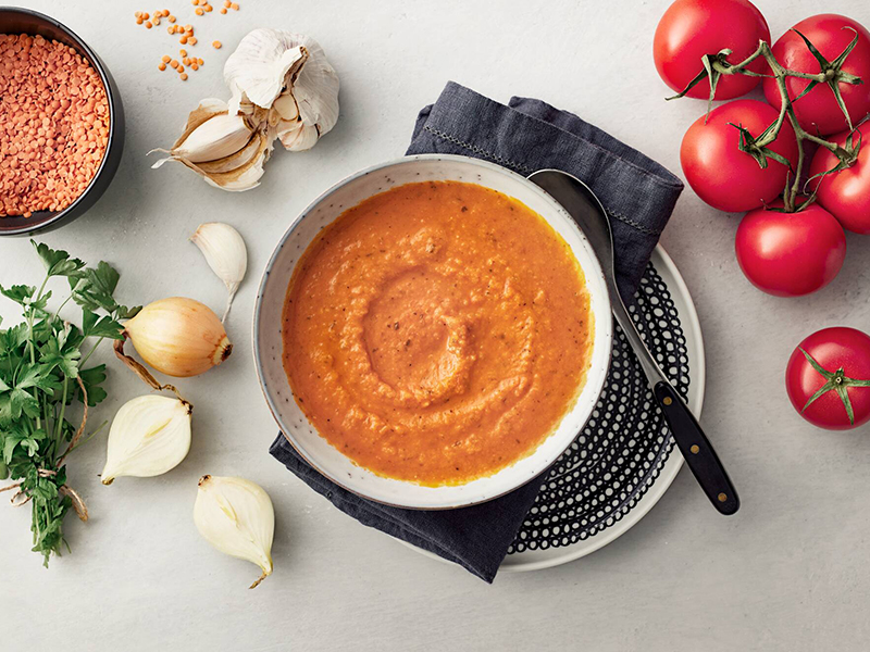 Kuvassa on kasvissosekeittoa lautasella ja pöydällä valkosipulia ja tomaattia