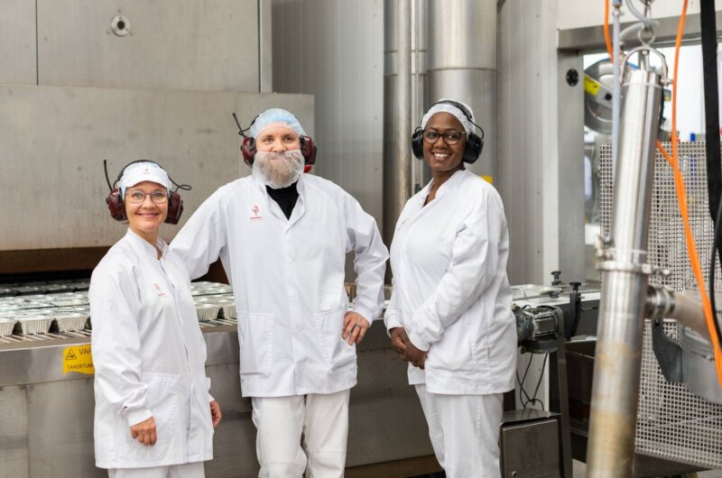 Kuvassa on kolme työntekijää suojavarusteissa tehtaalla.