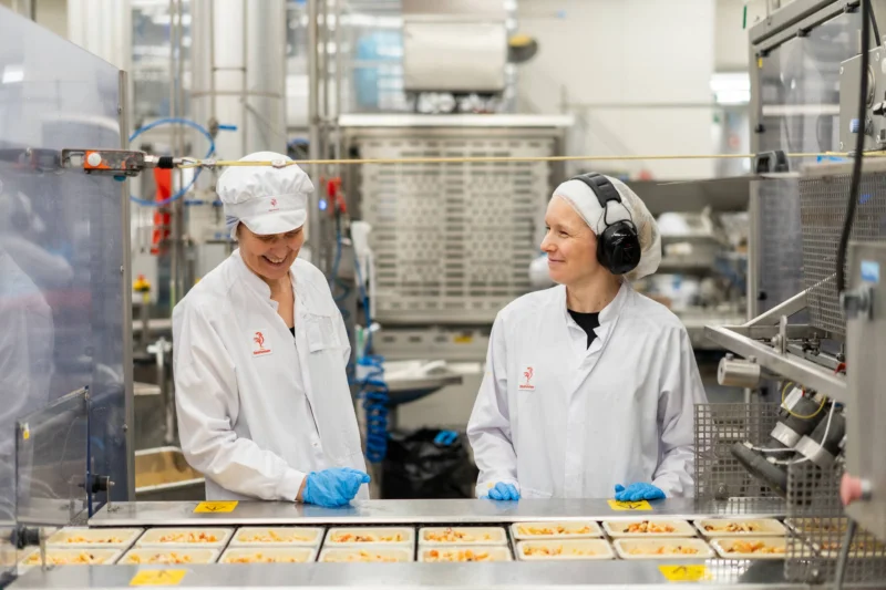 Kuvassa on kaksi henkilöä vieritysten katsomassa tehdaslinjaa.